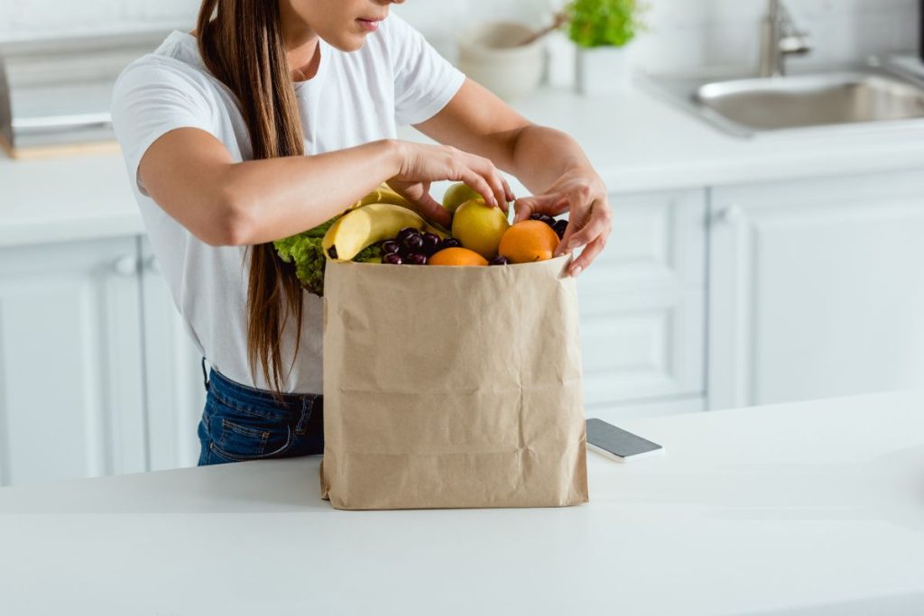 cropped photo of woman in white t-shirt and jeans looking through a paper bag of groceries in the kitchen
