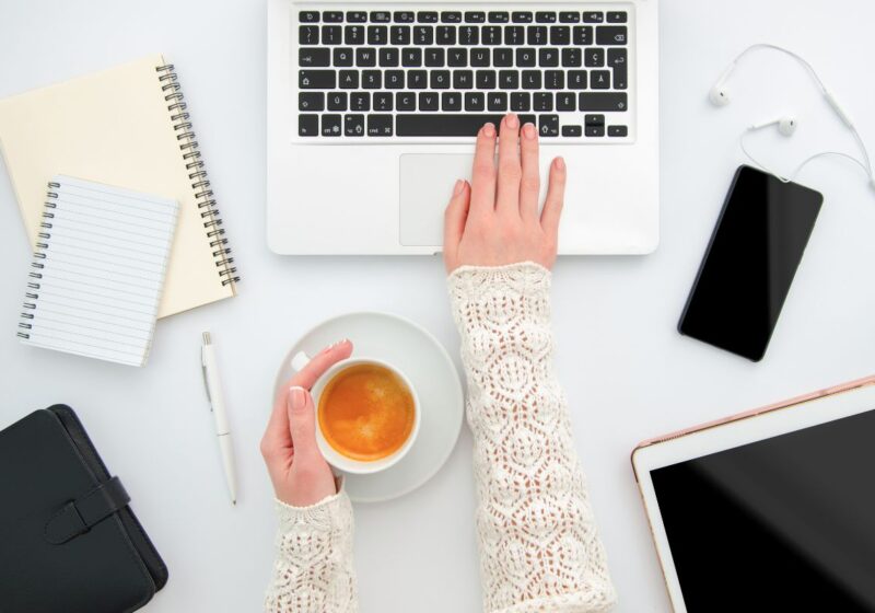 woman's hands, one holding a cup of coffee and one typing on laptop with planner, notebook, phone and tablet on the desk